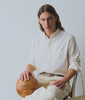 Man seated, wearing an off white oxford shirt and trousers, holding a soccer ball, against a white background