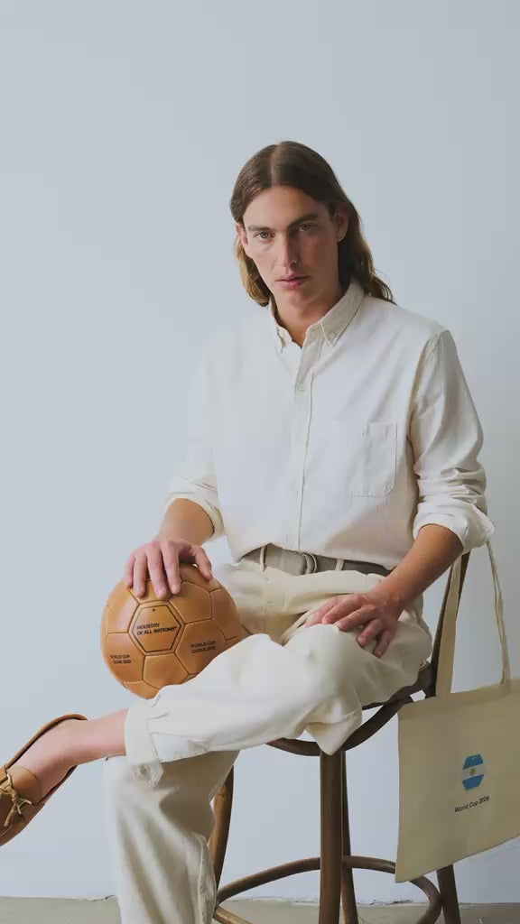 Model wearing undyed IOAN Gaucho Pants and an undyed IOAN Madras Shirt, sitting on a chair and playing with a leather soccer ball.
