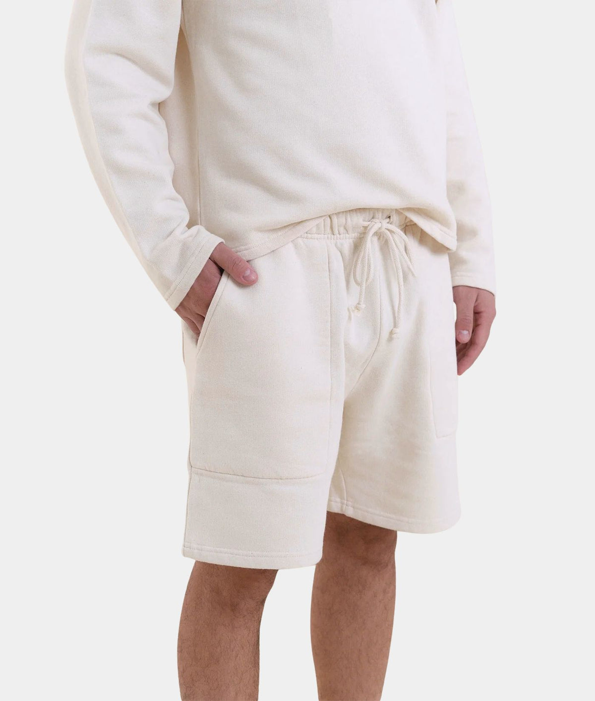 A person wearing cream-colored fleece shorts with a drawstring waist and a matching cream sweatshirt, shown from the chest down against a light gray studio background.