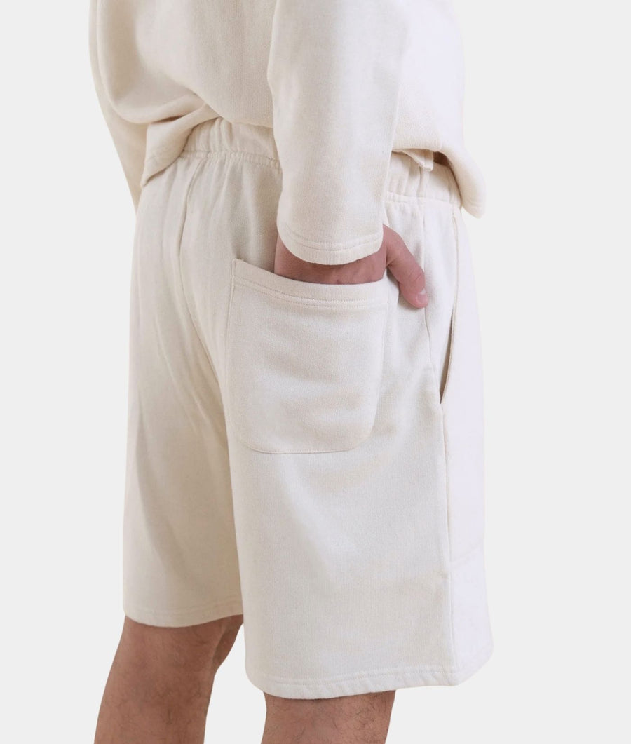 A close-up of a person wearing cream-colored cotton shorts with a large front patch pocket and a matching cream long-sleeve top, shown against a plain light background.