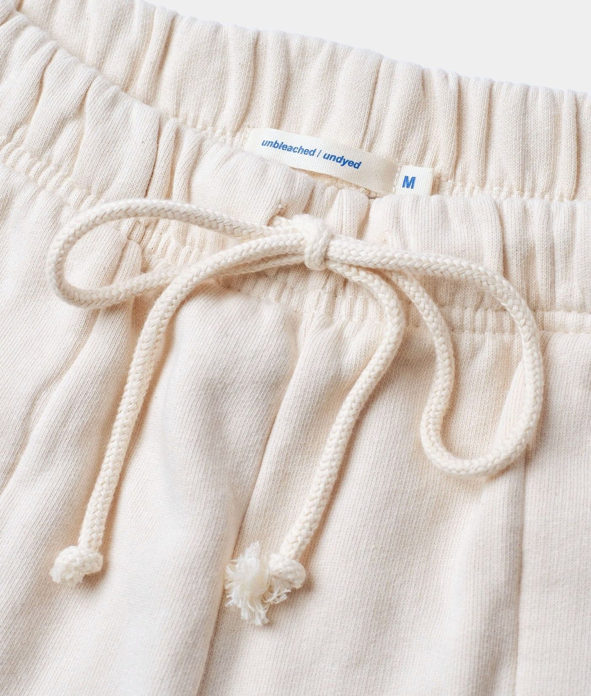 Close-up of cream-colored cotton lounge pants showing the elastic waistband, white drawstring, and a white brand label that reads 'unplugged / ungyed' with size 'M'.