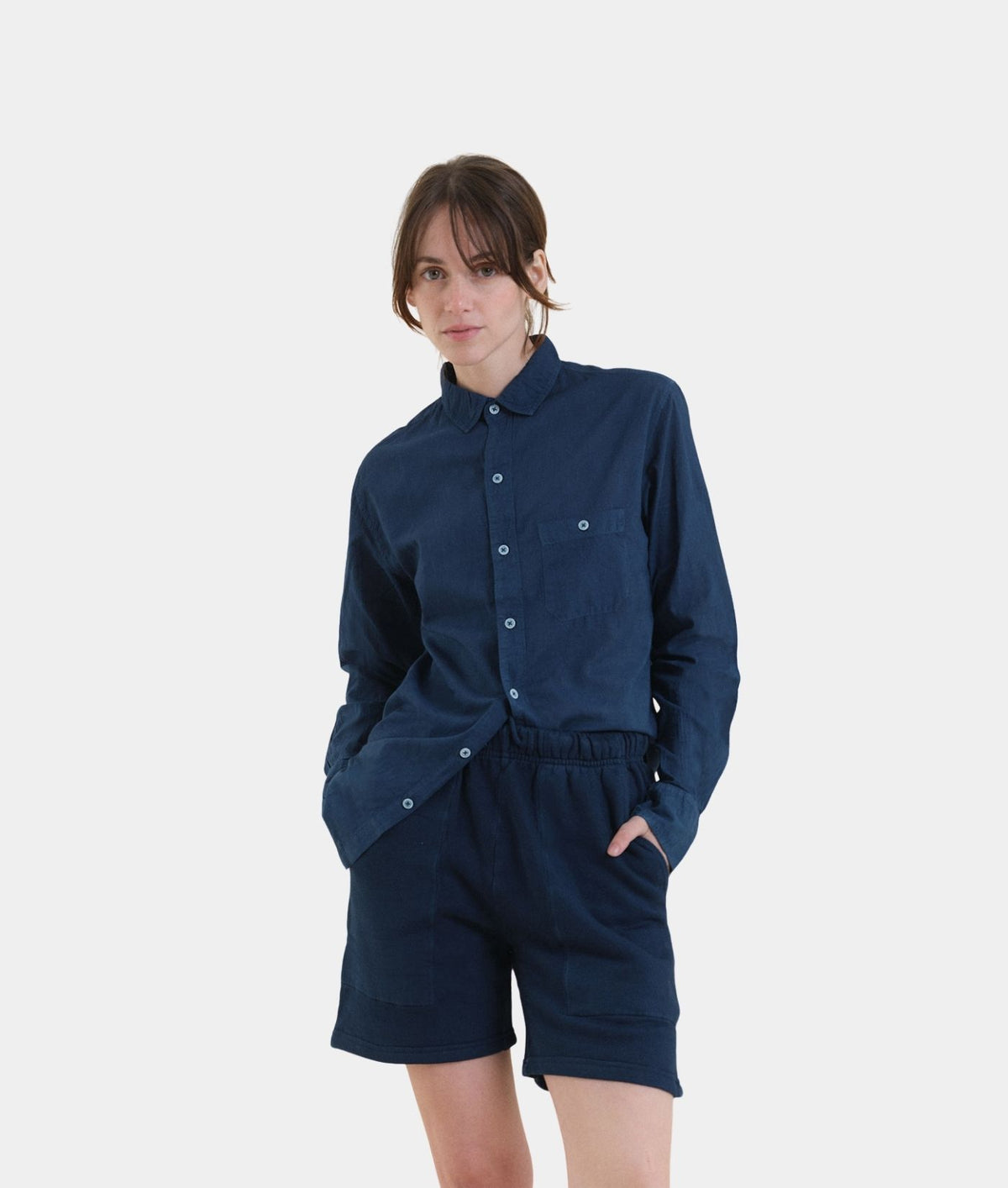 A woman modeling a dark navy blue matching set consisting of a long-sleeve button-up shirt and high-waisted shorts, photographed against a solid light gray background.