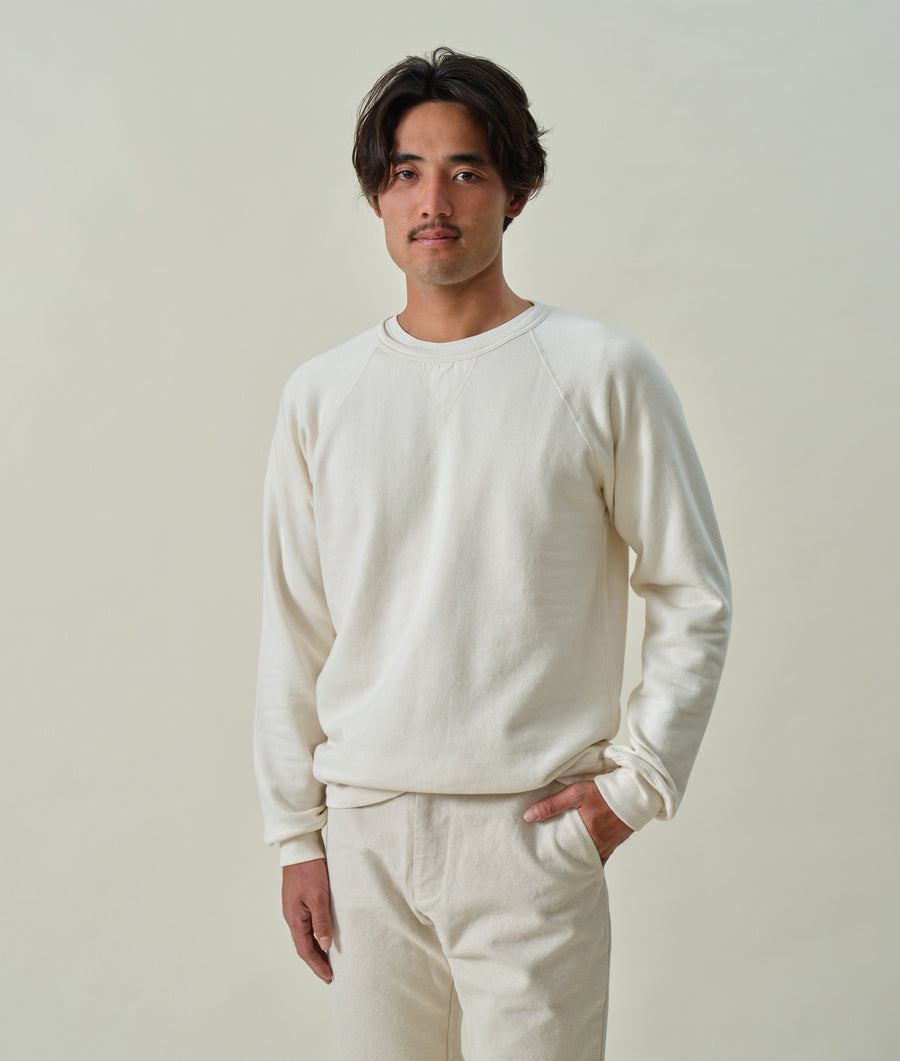 A young man wearing a long-sleeved, off-white organic cotton sweatshirt in a portrait shot against a light background - Industry of All Nations