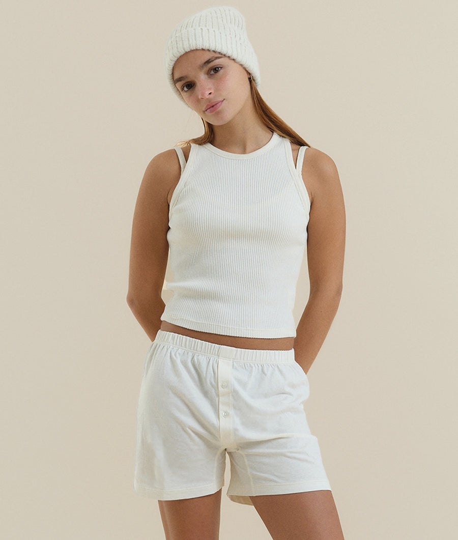 A young woman wearing a white ribbed tank top and matching white shorts, standing against a neutral background - Industry of All Nations