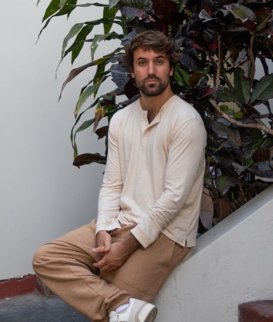 A bearded man sits on a white wall outdoors, wearing an IOAN Long Sleeve Henley Shirt in Caramel with tan pants.
