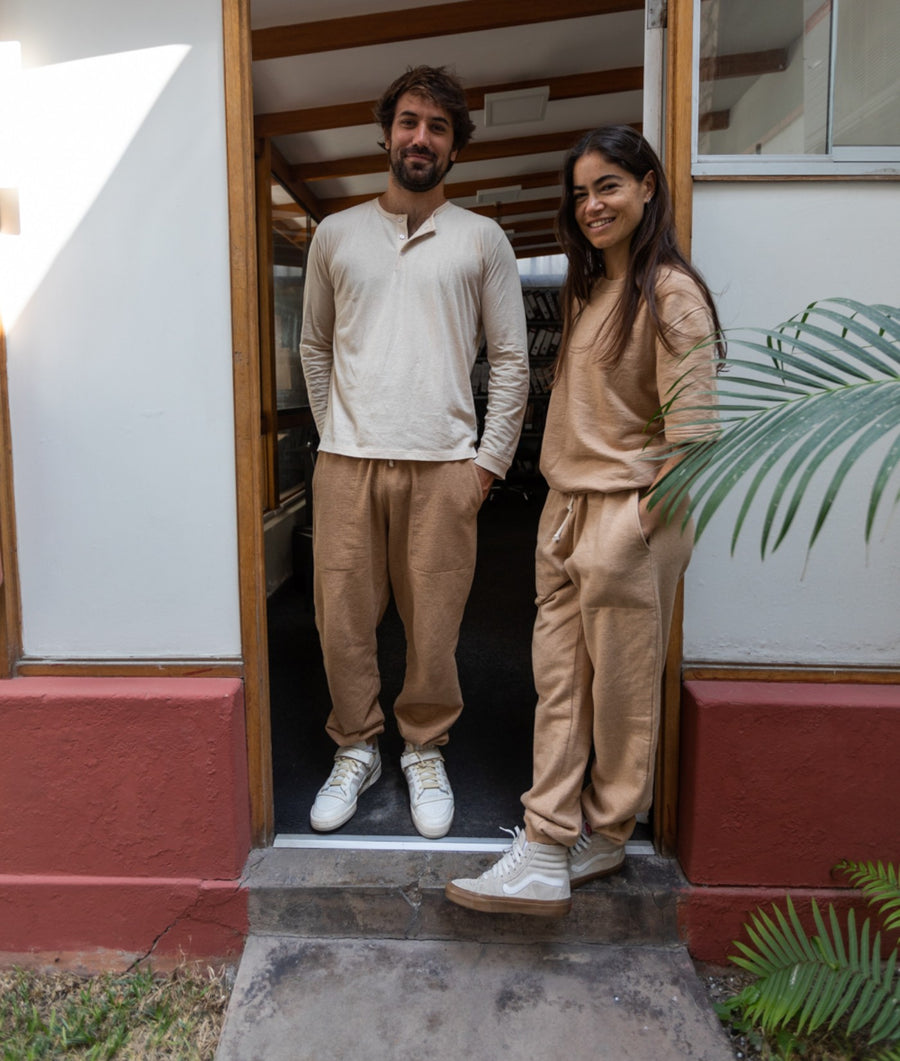 A man and a woman stand in a doorway wearing matching beige loungewear sets. The man wears a cream long-sleeve henley shirt and tan pants, while the woman wears a tan long-sleeve top and matching tan pants.