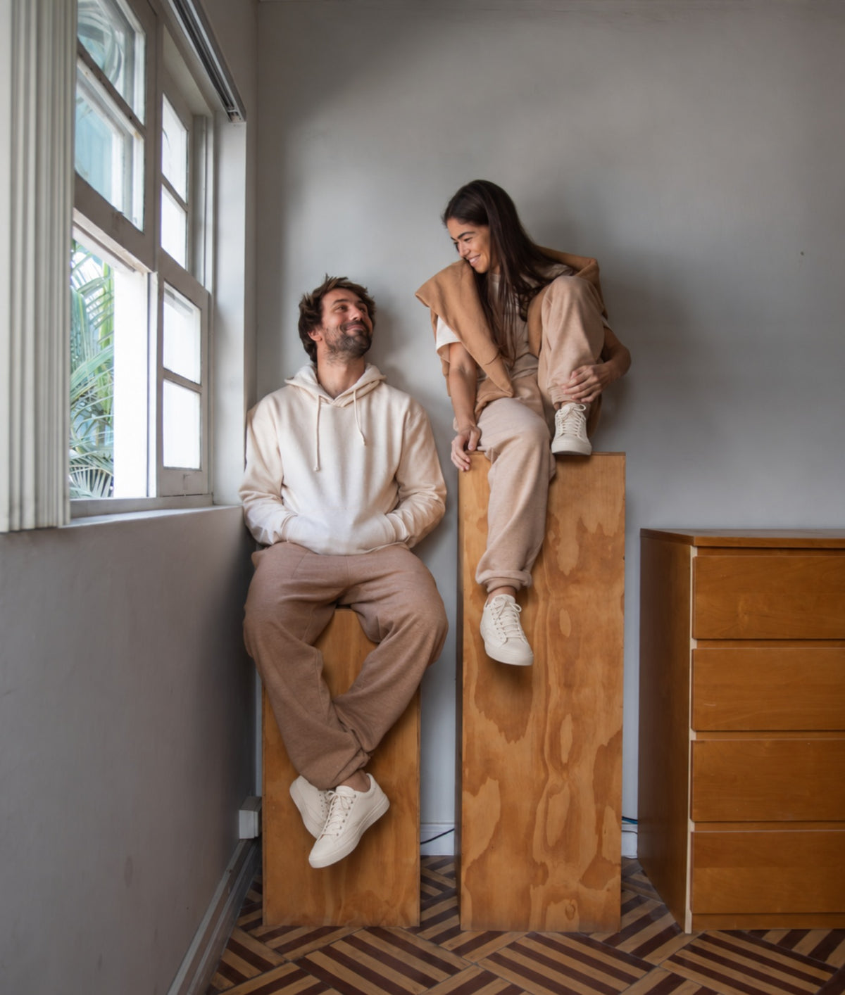 A man and a woman in matching beige and cream loungewear sit on wooden display blocks in a minimalist room with a window, gray walls, and a wood floor.