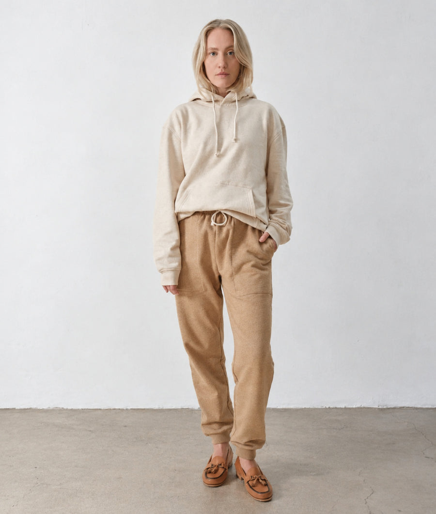 A woman stands against a plain white wall wearing a light beige hooded sweatshirt and tan drawstring pants, with brown slip-on shoes.