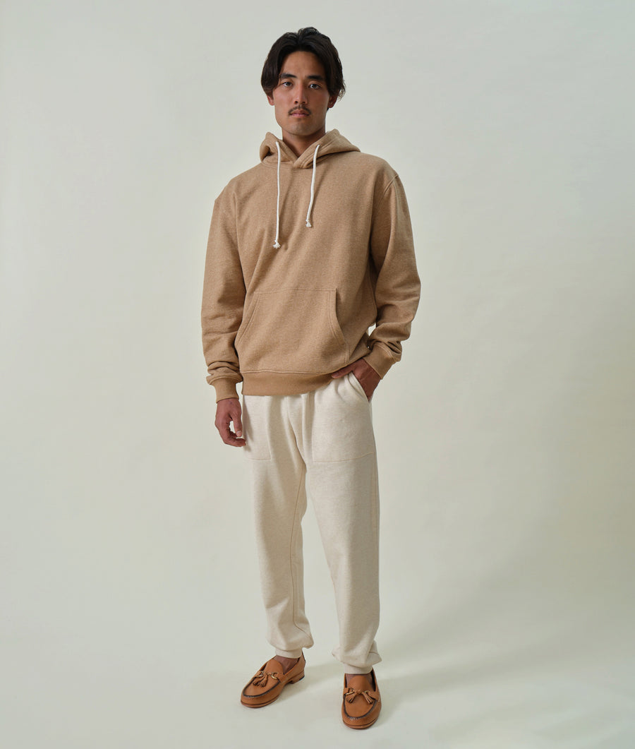 Man wearing a beige hooded sweatshirt and cream-colored sweatpants, standing in a plain setting - Industry of All Nations