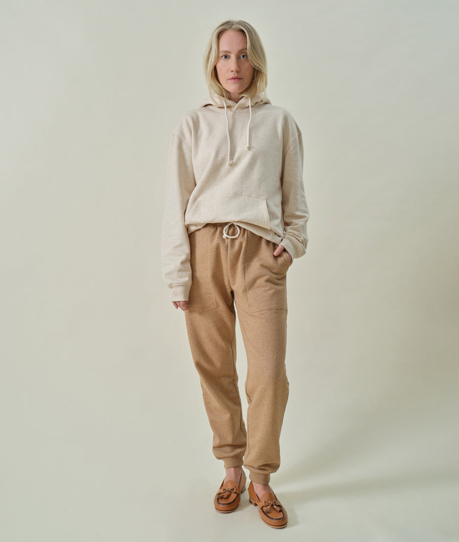 The image shows a woman wearing a beige hoodie and matching beige sweatpants, standing against a light background. - Industry of All Nations